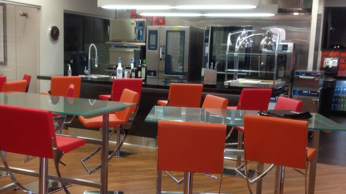 Demo kitchen with a black top counter, sink, commercial appliances and deep orange high chairs arranged around two glass tables